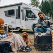 一對男女在森林中的露營地旁，坐在露營椅與地墊上喝咖啡、閱讀與放鬆，前方有一隻棕色捲毛狗陪伴，背景是一台停放在樹林邊的白色露營車，呈現悠閒的戶外生活氛圍。