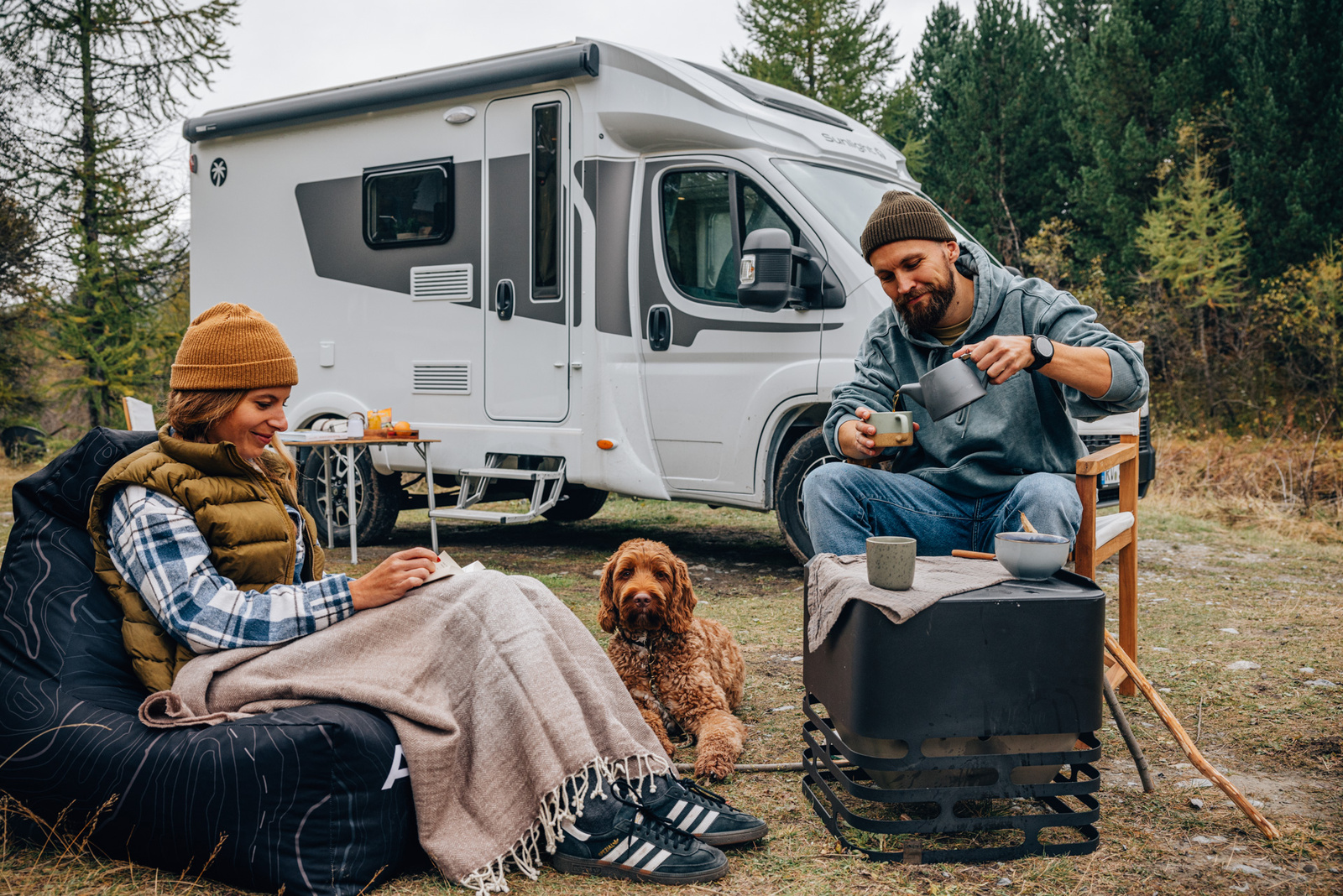 一對男女在森林中的露營地旁，坐在露營椅與地墊上喝咖啡、閱讀與放鬆，前方有一隻棕色捲毛狗陪伴，背景是一台停放在樹林邊的白色露營車，呈現悠閒的戶外生活氛圍。