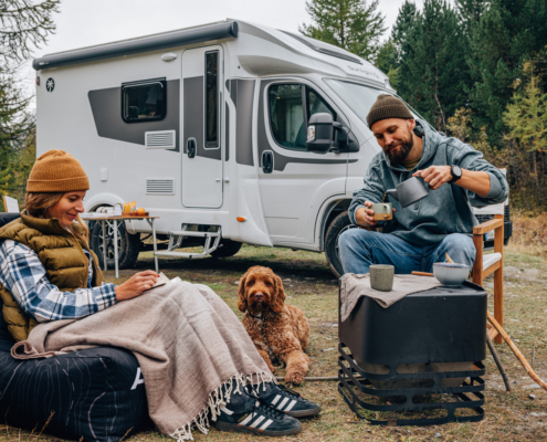 一對男女在森林中的露營地旁，坐在露營椅與地墊上喝咖啡、閱讀與放鬆，前方有一隻棕色捲毛狗陪伴，背景是一台停放在樹林邊的白色露營車，呈現悠閒的戶外生活氛圍。