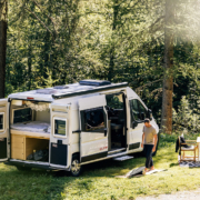 森林中的白色露營車，後門與側門敞開展示車內床鋪與收納設計，兩位旅人在露營車旁的戶外桌椅休息，呈現自駕露營與車宿生活情境