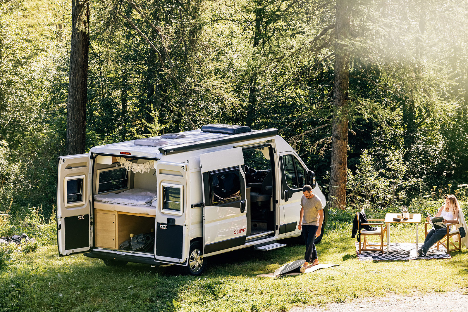 森林中的白色露營車，後門與側門敞開展示車內床鋪與收納設計，兩位旅人在露營車旁的戶外桌椅休息，呈現自駕露營與車宿生活情境