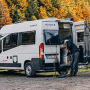 Sunlight Cliff 露營車停靠在秋季森林湖畔，車主於車尾整理露營裝備與自行車