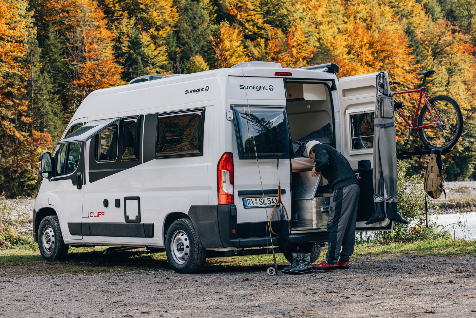 Sunlight Cliff 露營車停靠在秋季森林湖畔，車主於車尾整理露營裝備與自行車